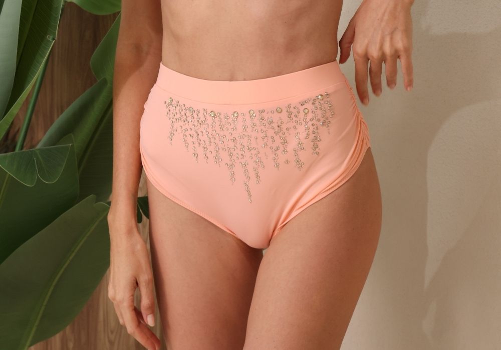 Person wearing peach high-waisted bikini bottoms with embellishments against a neutral background.